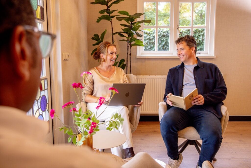 Personen die een training volgen of vergaderen bij vergaderlocatie Ruimte voor Inzicht in Oisterwijk. Ze zitten op comfortabele stoelen in een ruimte met veel licht en uitzicht op het groen.