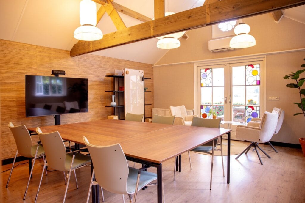 Opstelling voor 6 personen bij vergaderlocatie Ruimte voor Inzicht met comfortabele stoelen, tv, verrijdbare tafels en glasbord. Veel licht en openslaande deuren naar buiten