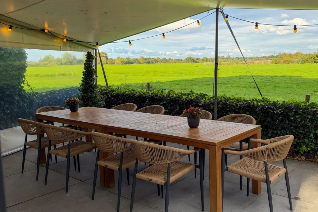 Overdekt terras met twee teak houten tafels en 10 stoelen met uitzicht op het groen bij vergaderlocatie Ruimte voor Inzicht in Oisterwijk